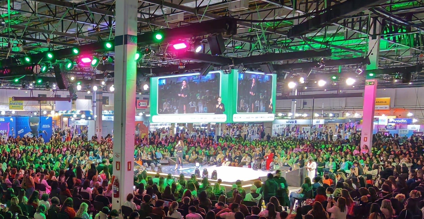 Telões suspensos sobre a plenária lotada de um summit, com a palestrante em destaque e o intérprete de Libras em quadro próprio ao lado