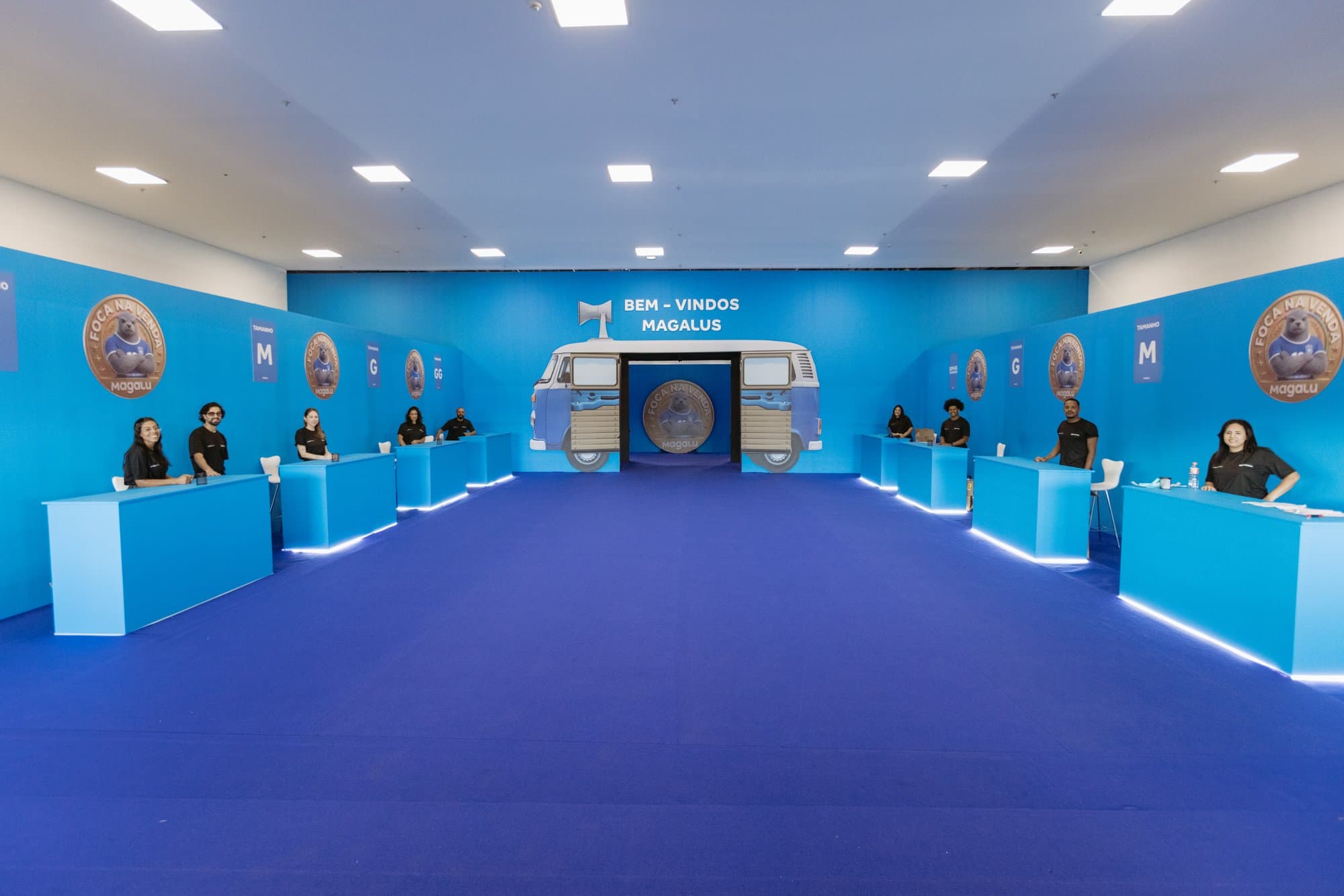 Sala de credenciamento com balcões iluminados e painéis com a identidade do evento, e uma cenografia de boas-vindas ao fundo