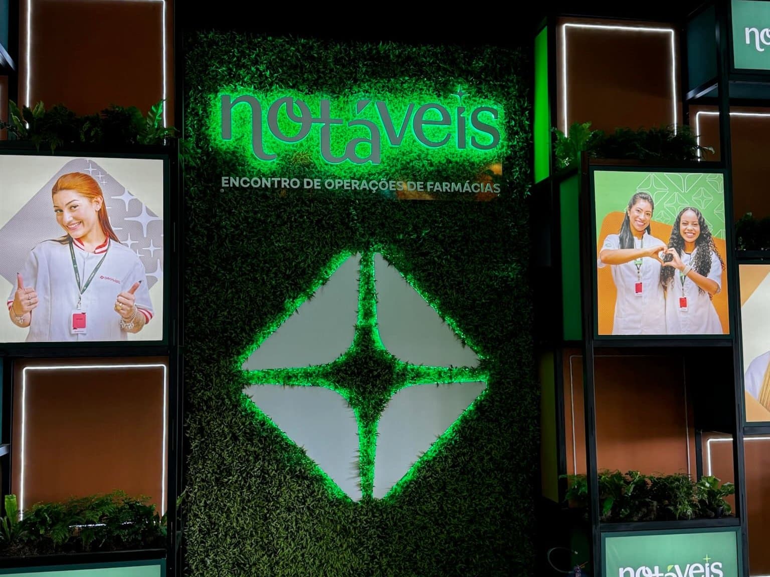 Parede instagramável com o logo do evento iluminado sobre jardim vertical, módulos com fotos de colaboradores e o símbolo da marca em LED verde
