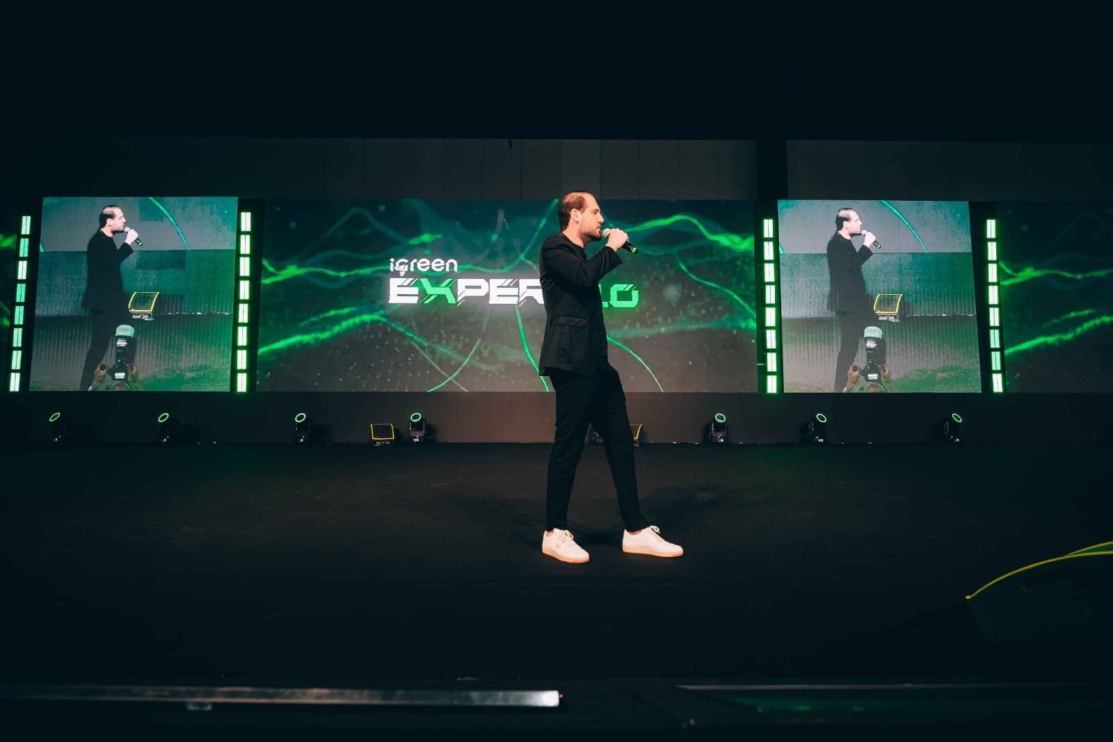 Palestrante no palco diante do telão de LED com o KV do evento em verde e dois telões laterais com a imagem ao vivo