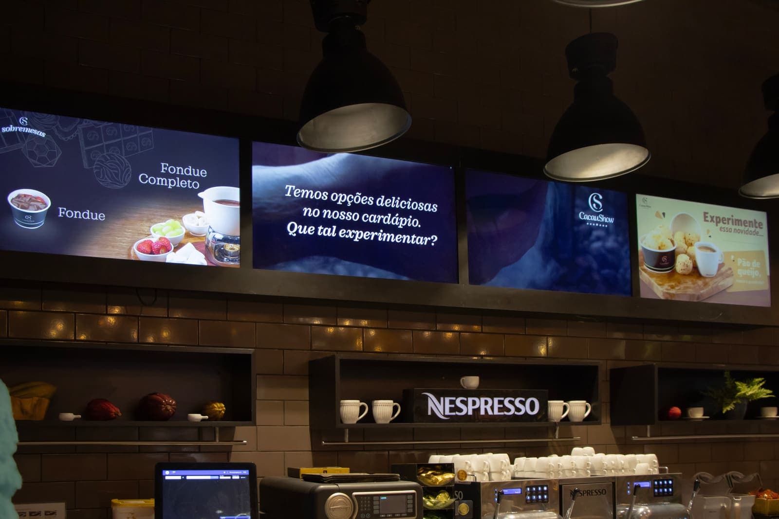 Menu board digital da Cacau Show: quatro telas sobre o balcão exibindo sobremesas e chamadas do cardápio