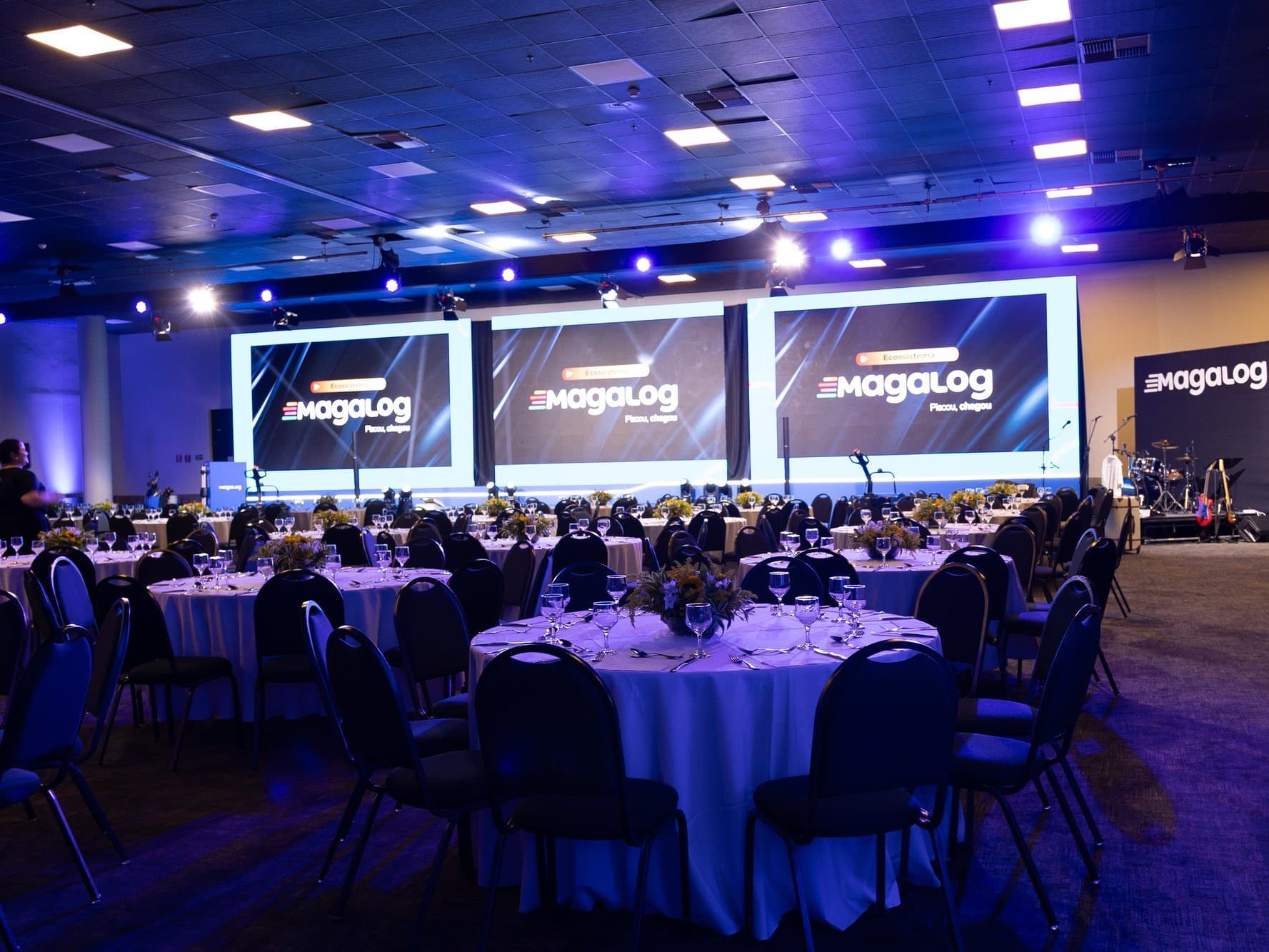 Salão de jantar com mesas redondas postas diante de três telões de LED com o KV do evento