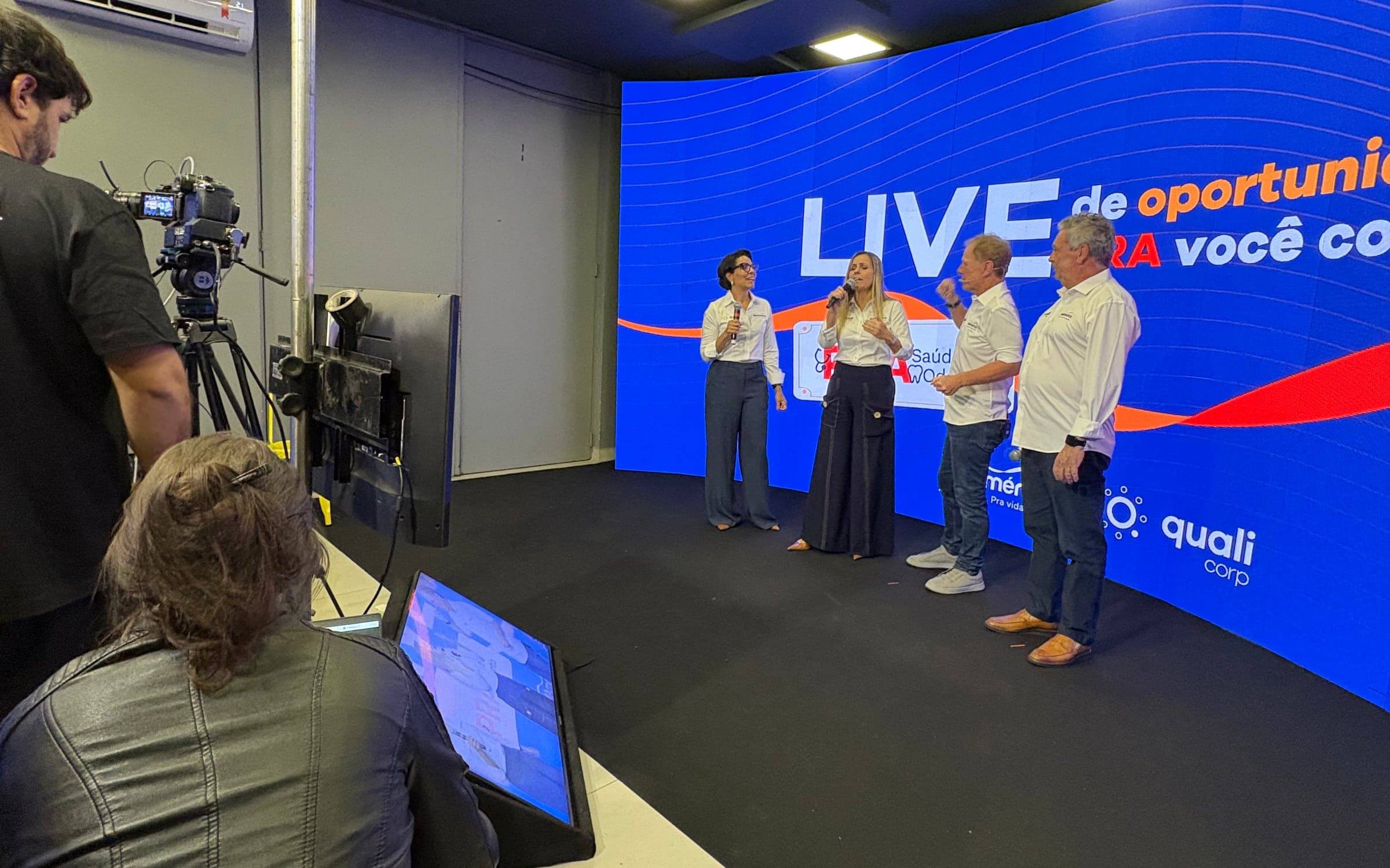 Live corporativa em estúdio, com quatro apresentadores diante do painel de LED curvo e a câmera em primeiro plano