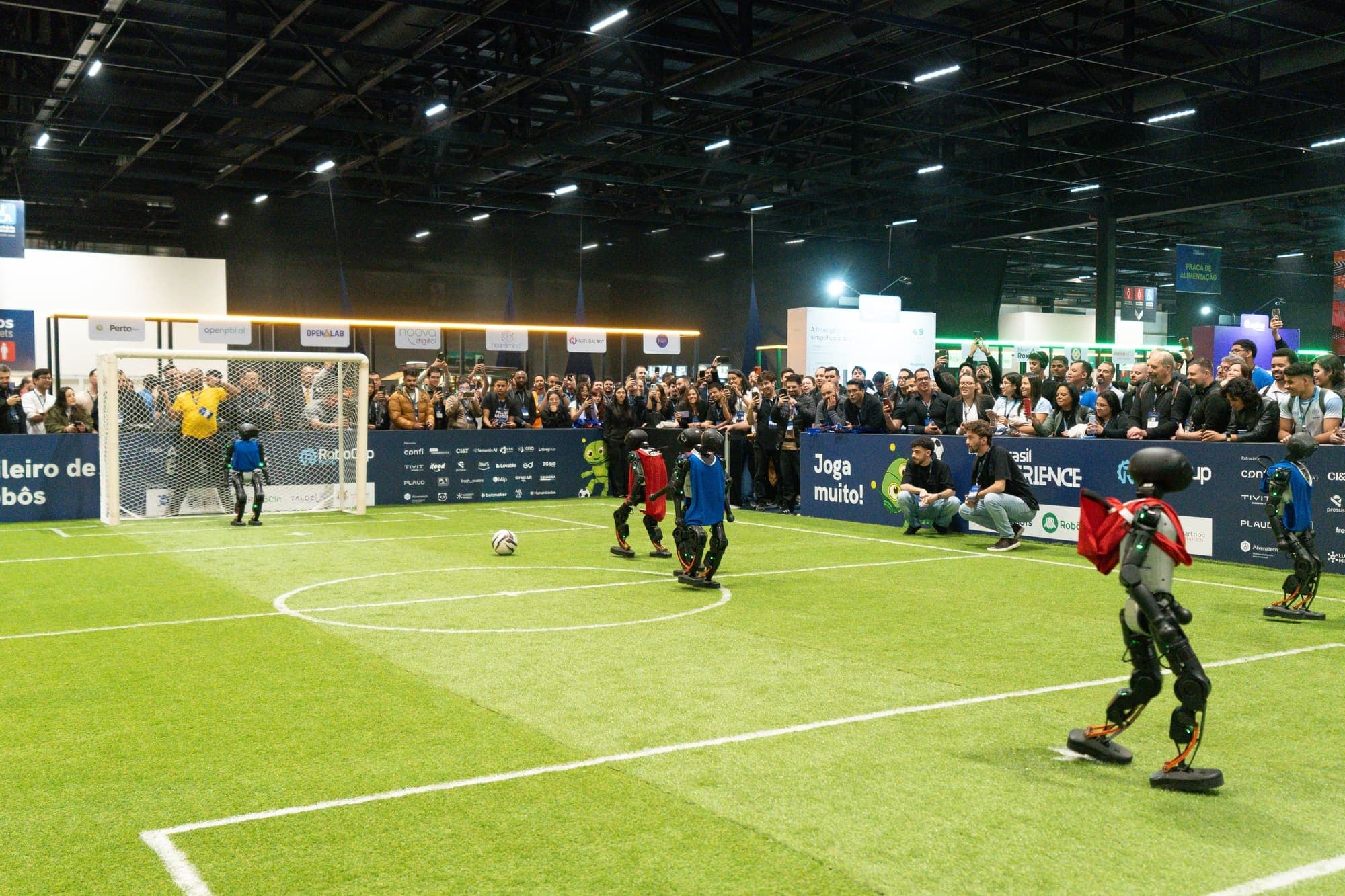 Arena com campo de grama sintética e traves, onde robôs disputam uma partida diante do público