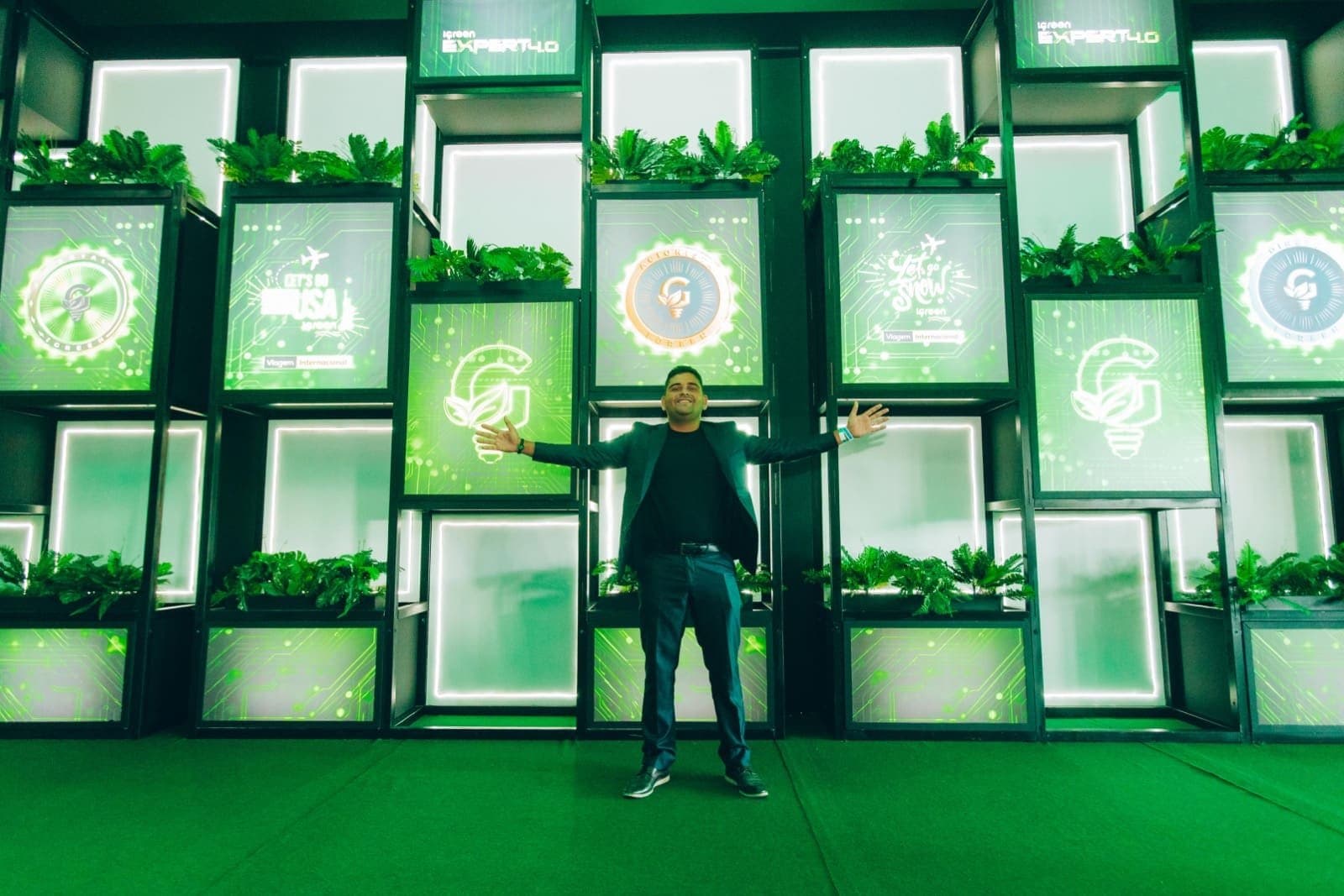 Parede instagramável de módulos iluminados com plantas e ícones do evento, com um participante posando de braços abertos