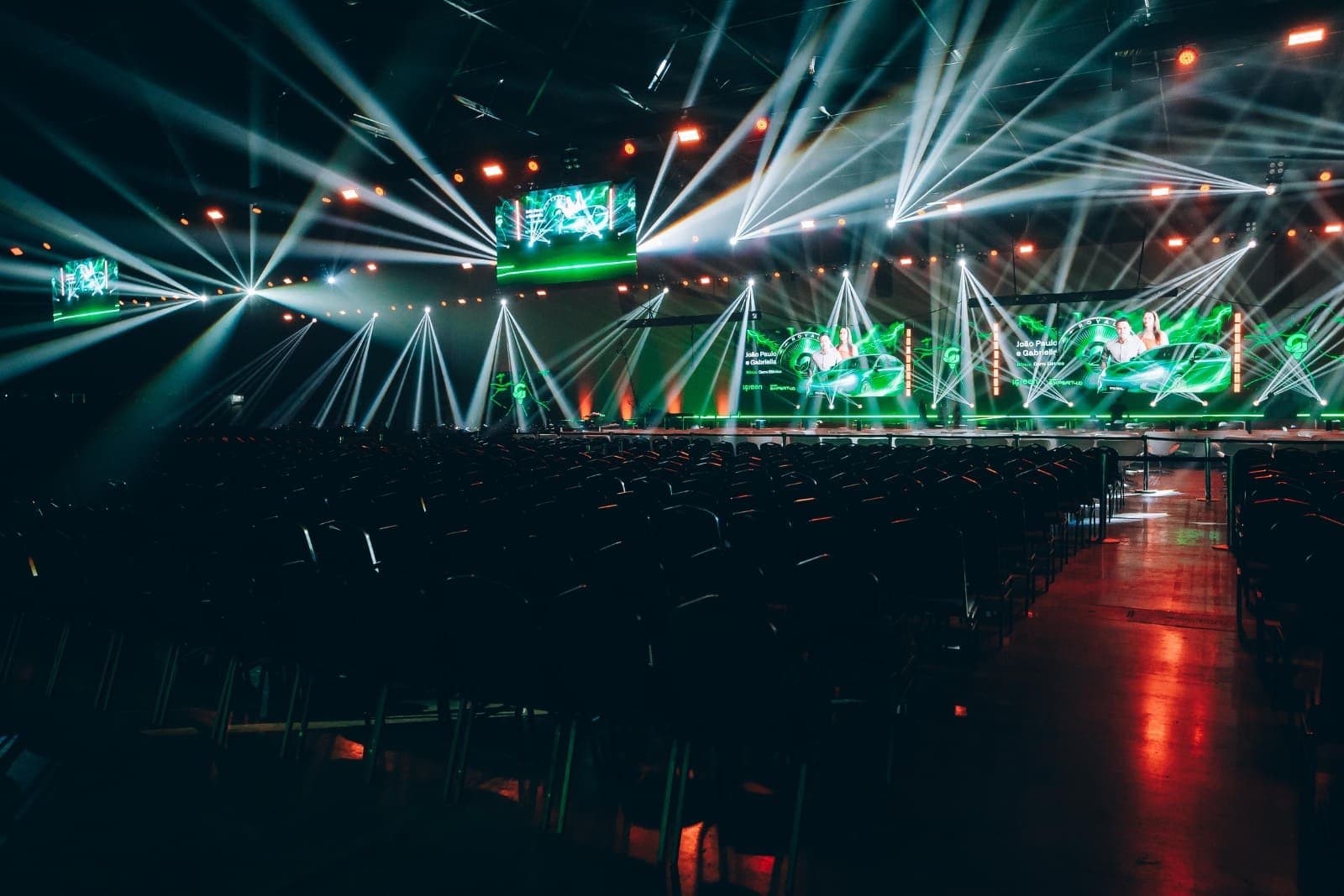 Plenária vista do fundo, com o palco iluminado por feixes de luz, painéis de LED e as cadeiras prontas para o público
