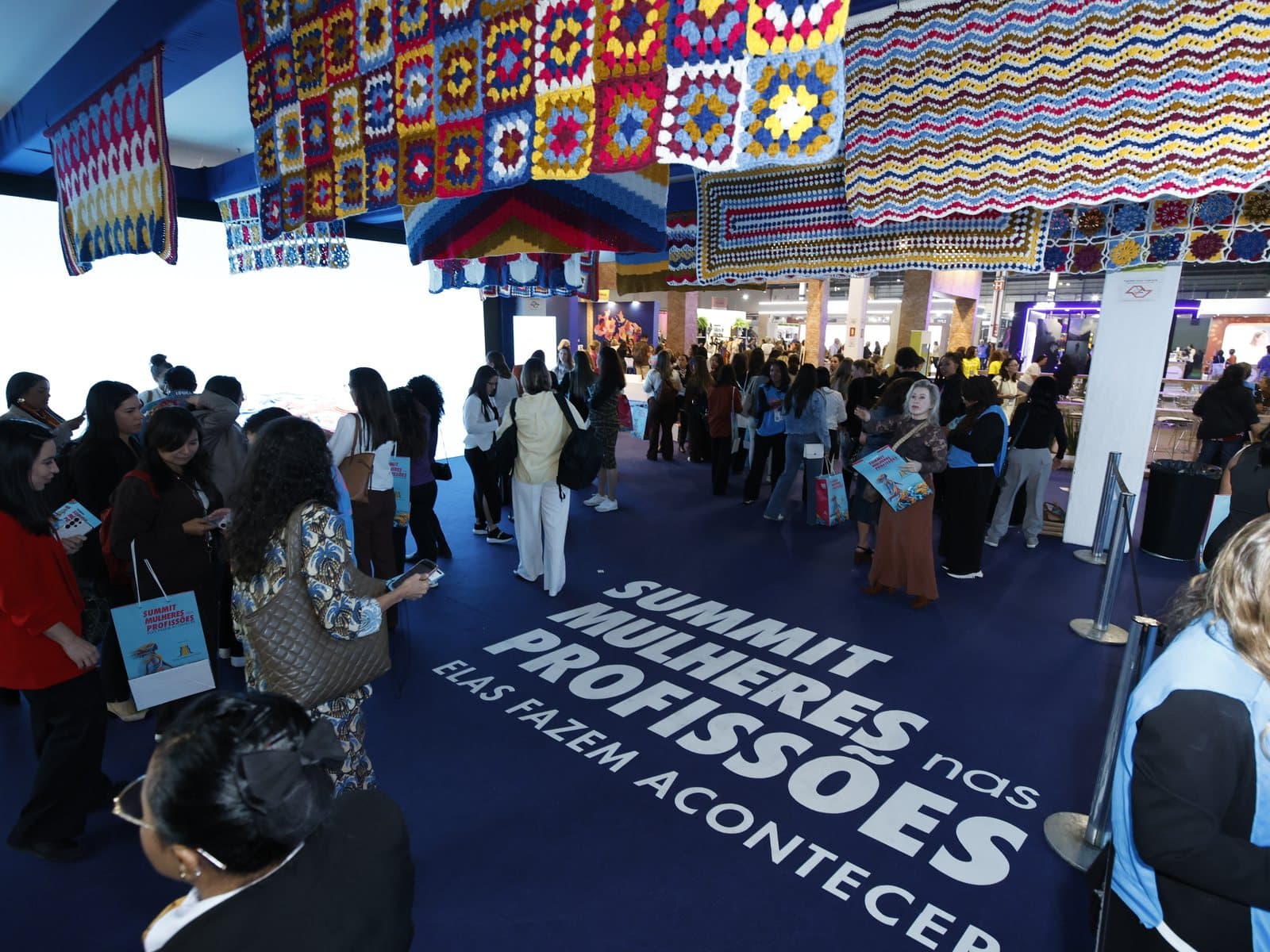 Público entrando no pavilhão sobre o tapete com o nome do evento, sob uma cobertura de crochê colorido